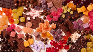 A flatlay image of a variety of KIVA confections on a beige background. There is a mixture of chocolate bars, little chocolate bite spheres, square fruit chews, diamond shaped mints, circular and diamond shaped gummies dusted in sugar and some gummies without sugar dustings.